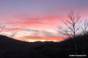 In the foreground some hills and vegetation with a tree silhouetted against a multi-colored sunset of yellows, orange, pink, and purple with scattered pink clouds.
