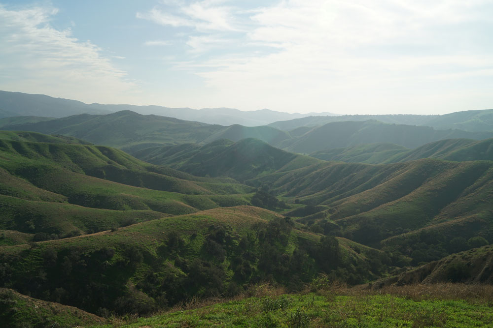 A view of multiple canyons and ridgelines in green and slightly brown shades with a hazy sky with medium blues and dusty white wispy clouds.