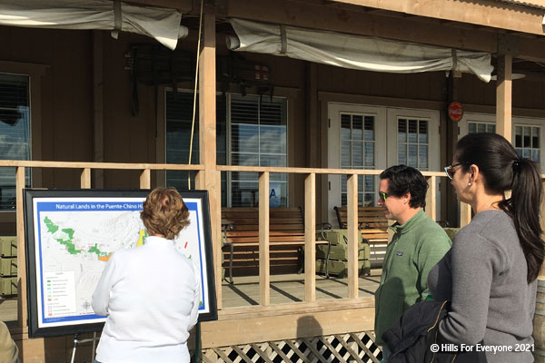 A woman in a white shirt explains through the use of a large map to two individuals, a man and a woman, the protected and unprotected landscape with a largely wooden building in the background.