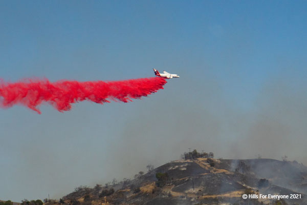 A white plane with a red tail drops bright red fire retardant on smoking black hills with charred vegetation against a dusty blue sky.