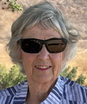 A woman smiles for the camera. She is wearing a blue and white striped shirt, sunglasses, and has green and brown vegetation behind her.