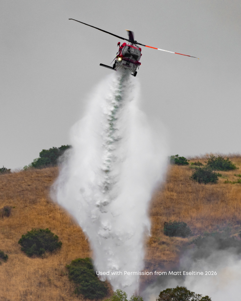 A helicopter making a water drop on smoldering hillsides.